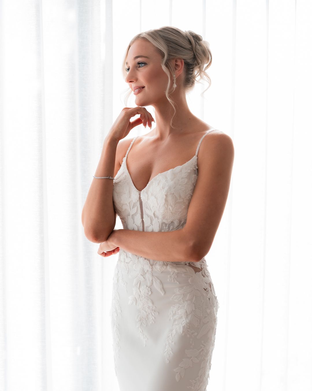 Elegant bride in a lace spaghetti strap wedding gown posing gracefully by sheer white curtains.