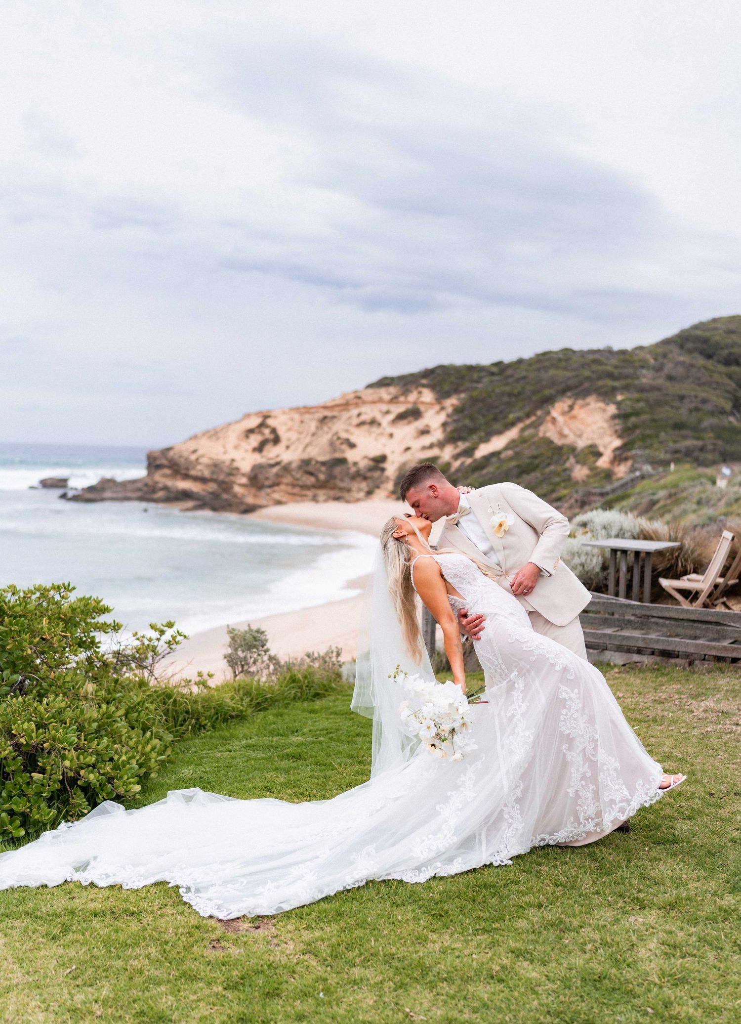 A couple in wedding attire embraces on a grassy cliff overlooking a dramatic coastal beach landscape.