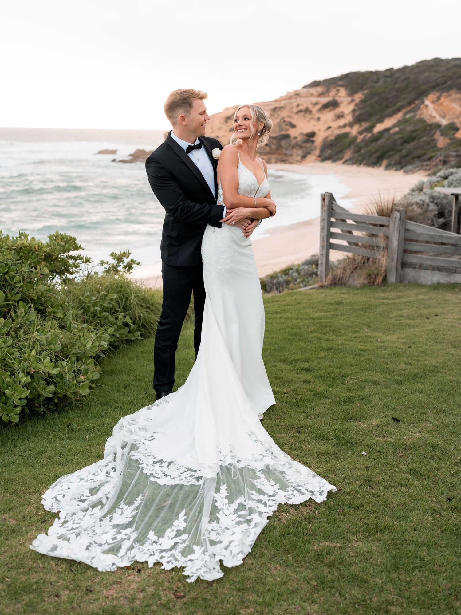 Bride in lace train gown and groom in tuxedo embrace on coastal lawn with sandy beach and cliffs backdrop.