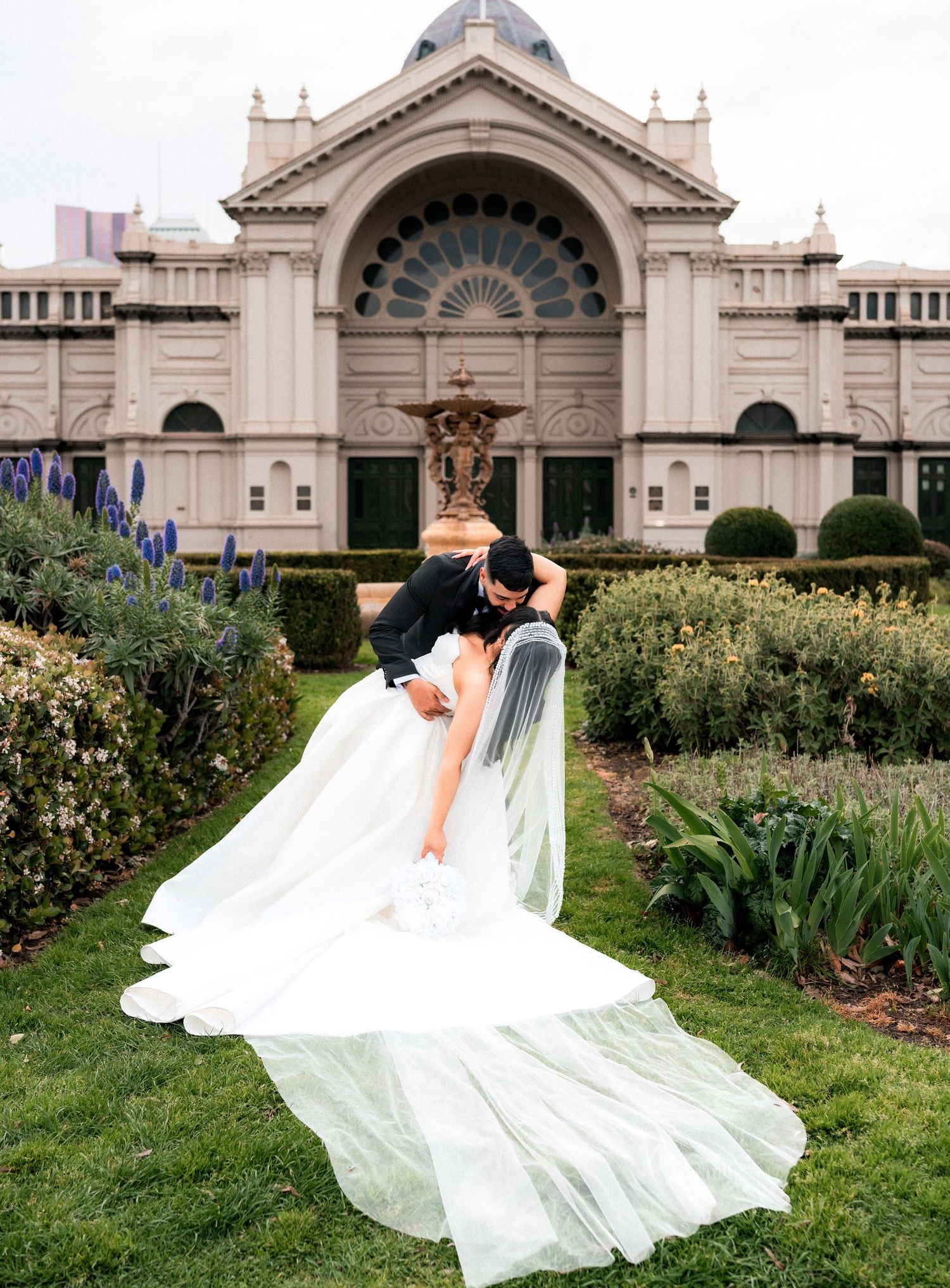 A flowing white wedding dress and veil create an elegant silhouette against the backdrop of a grand cathedral garden.