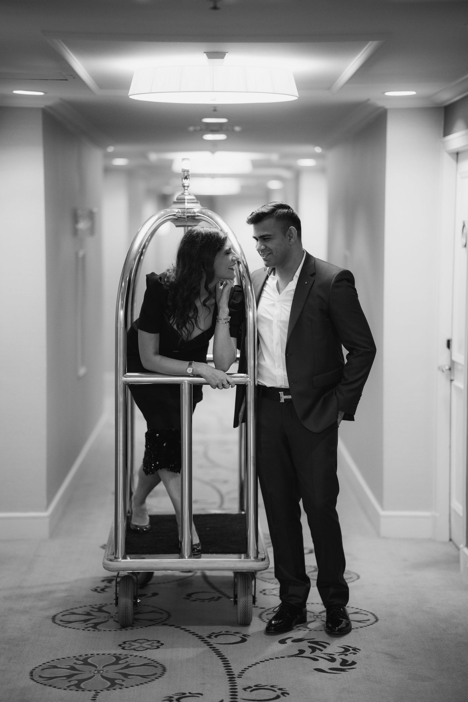 A couple stands in a hotel hallway next to an ornate luggage cart, captured in elegant black and white photography.