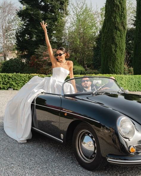 Classic black vintage Porsche 356 convertible with bride in white wedding dress posing joyfully in a garden setting.