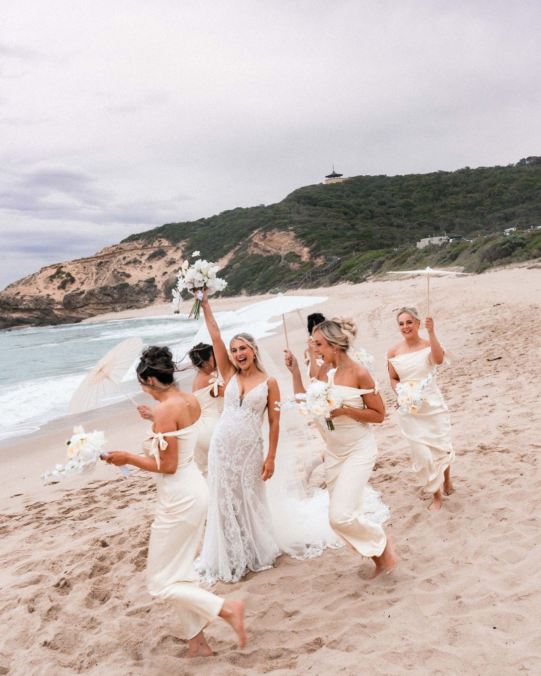 A group in white dresses playfully runs and poses together on a scenic beach with rocky cliffs in the background.