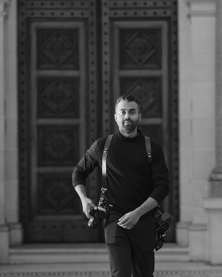 A black and white photo of a person in dark clothing with suspenders walking in front of a brick wall.