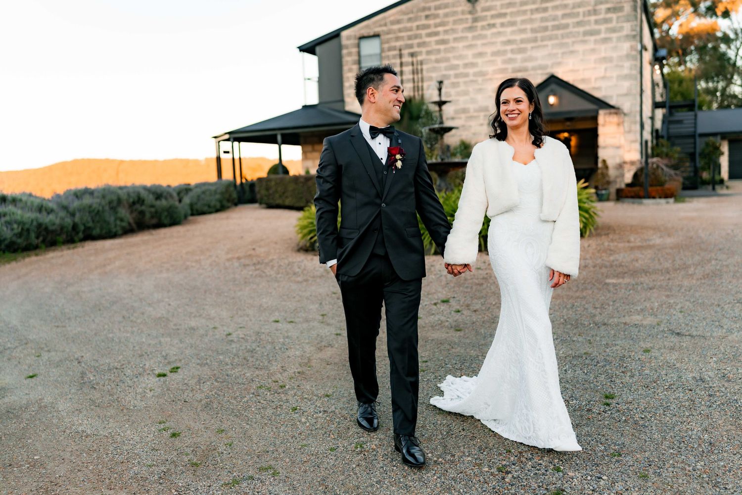 Couple in formal wedding attire walking together at sunset on a dirt path with mountains in the background.