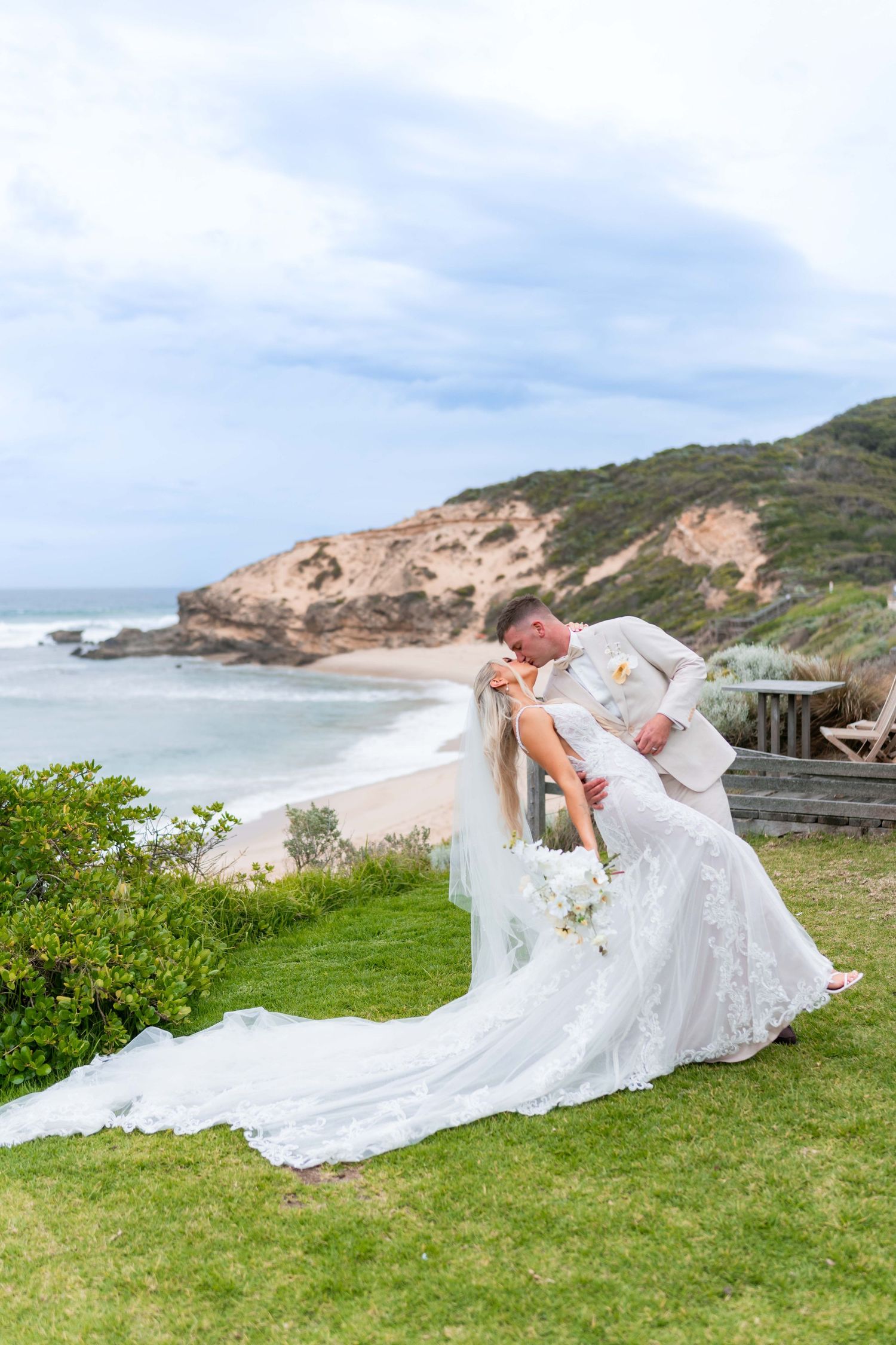 A bride in a flowing white dress poses dramatically on coastal cliffs overlooking a scenic beach landscape.