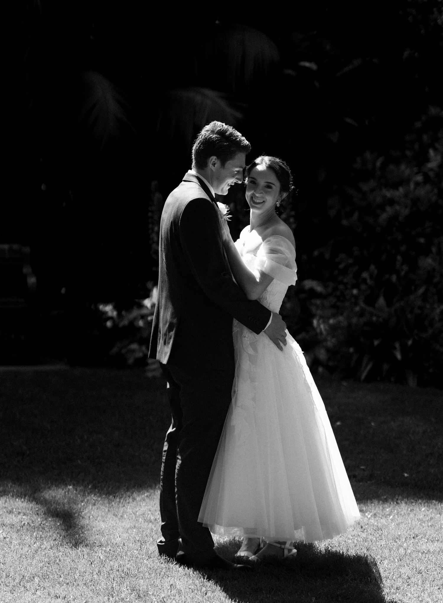 Black and white photo of a smiling bride in a white gown embracing her groom in a suit outdoors.