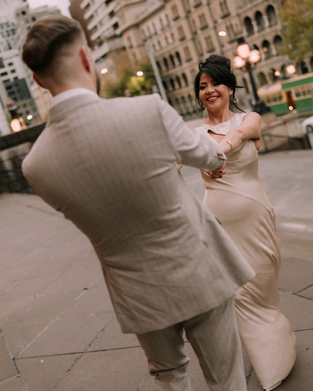Bride and groom dancing outdoors in elegant wedding attire on a city street at dusk.