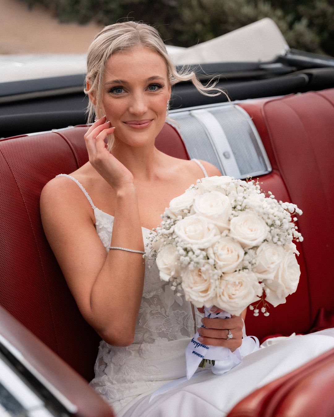 Radiant bride holding white rose bouquet seated in a red convertible car on her wedding day.