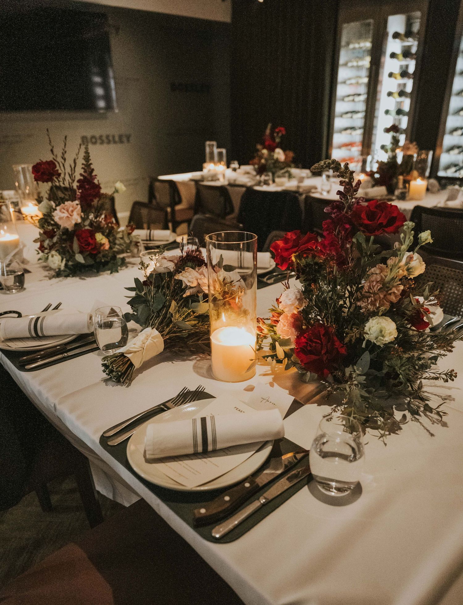 Elegant wedding reception table setting with moody lighting, red floral centerpieces and glowing candles on white linens.