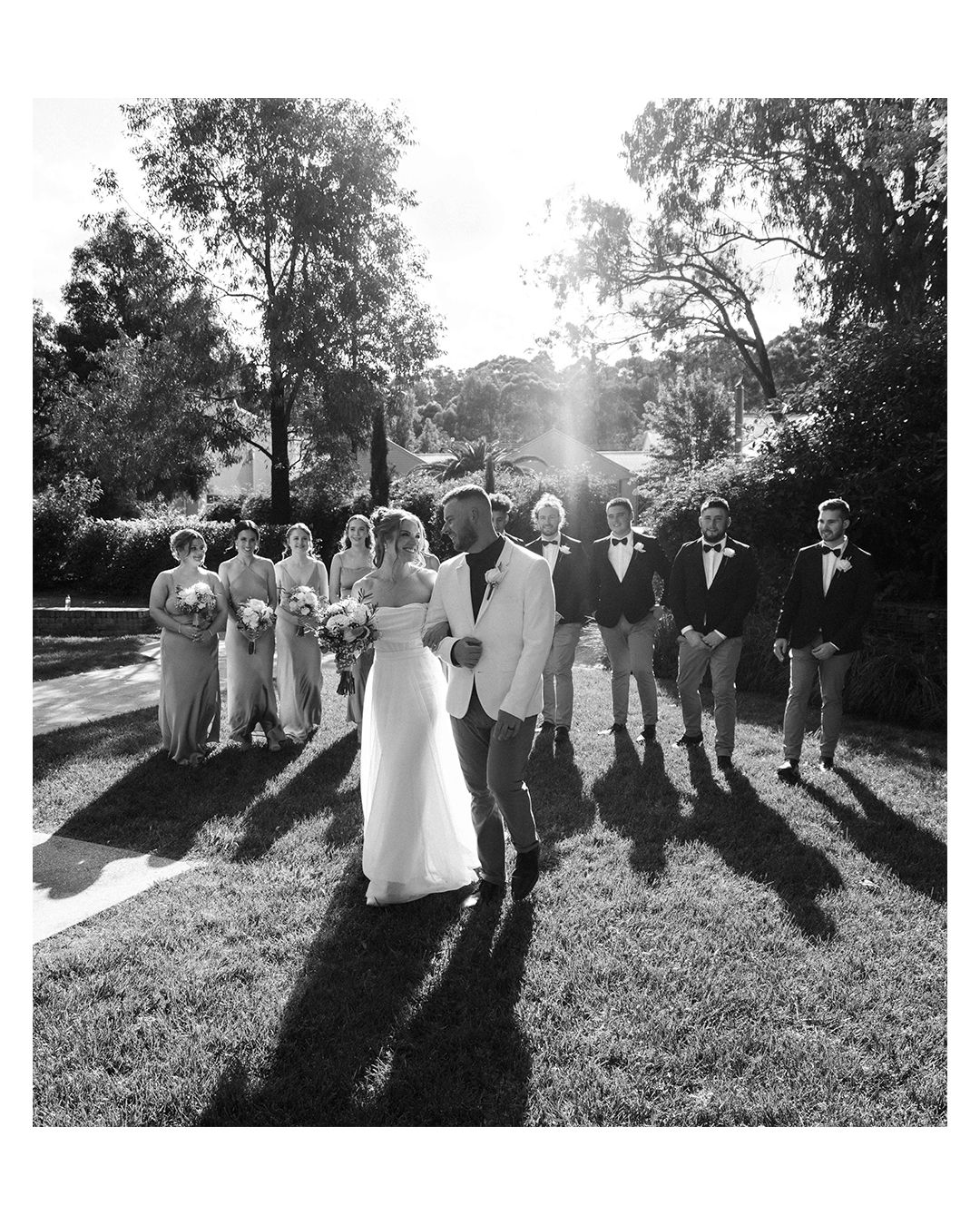 A wedding party stands in silhouette against sunlight casting long shadows across a grassy field in black and white.