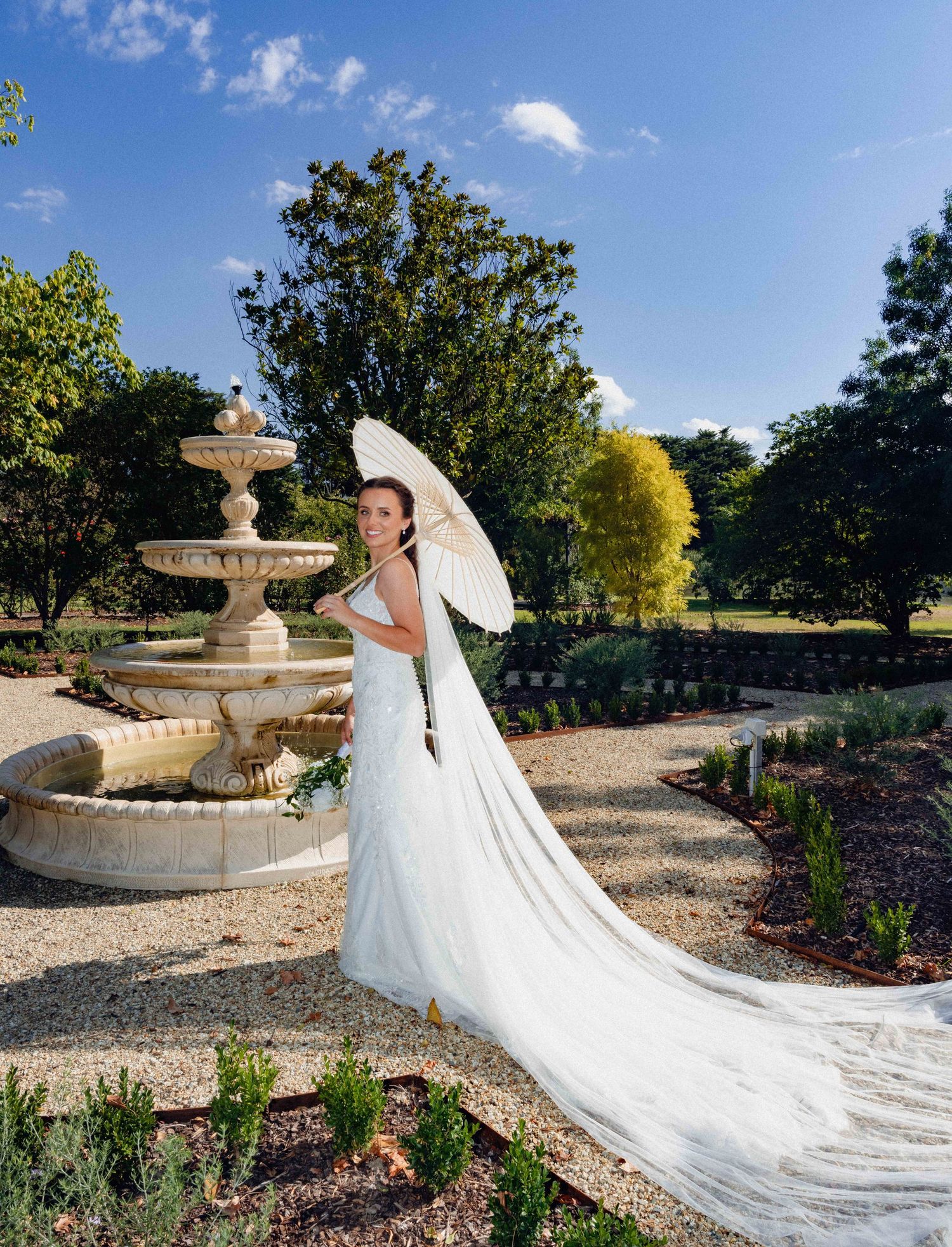 A flowing cathedral-length veil trails elegantly across a garden path near an ornate stone fountain.