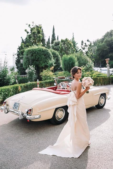 A vintage cream-colored convertible Mercedes-Benz roadster parked on a garden path with lush greenery in the background.