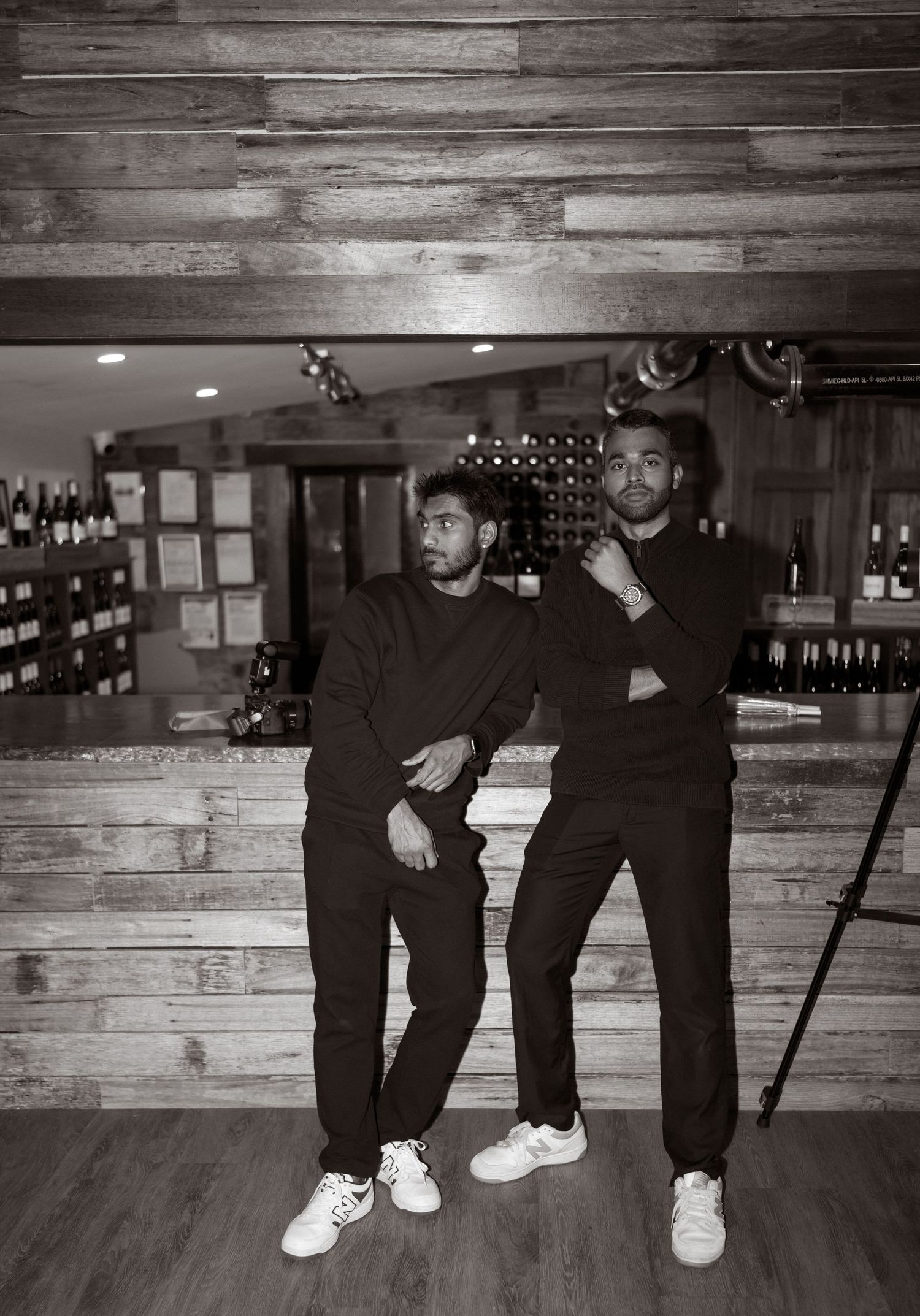 Two people in black outfits strike casual poses against a rustic wood wall backdrop in what appears to be a wine cellar.