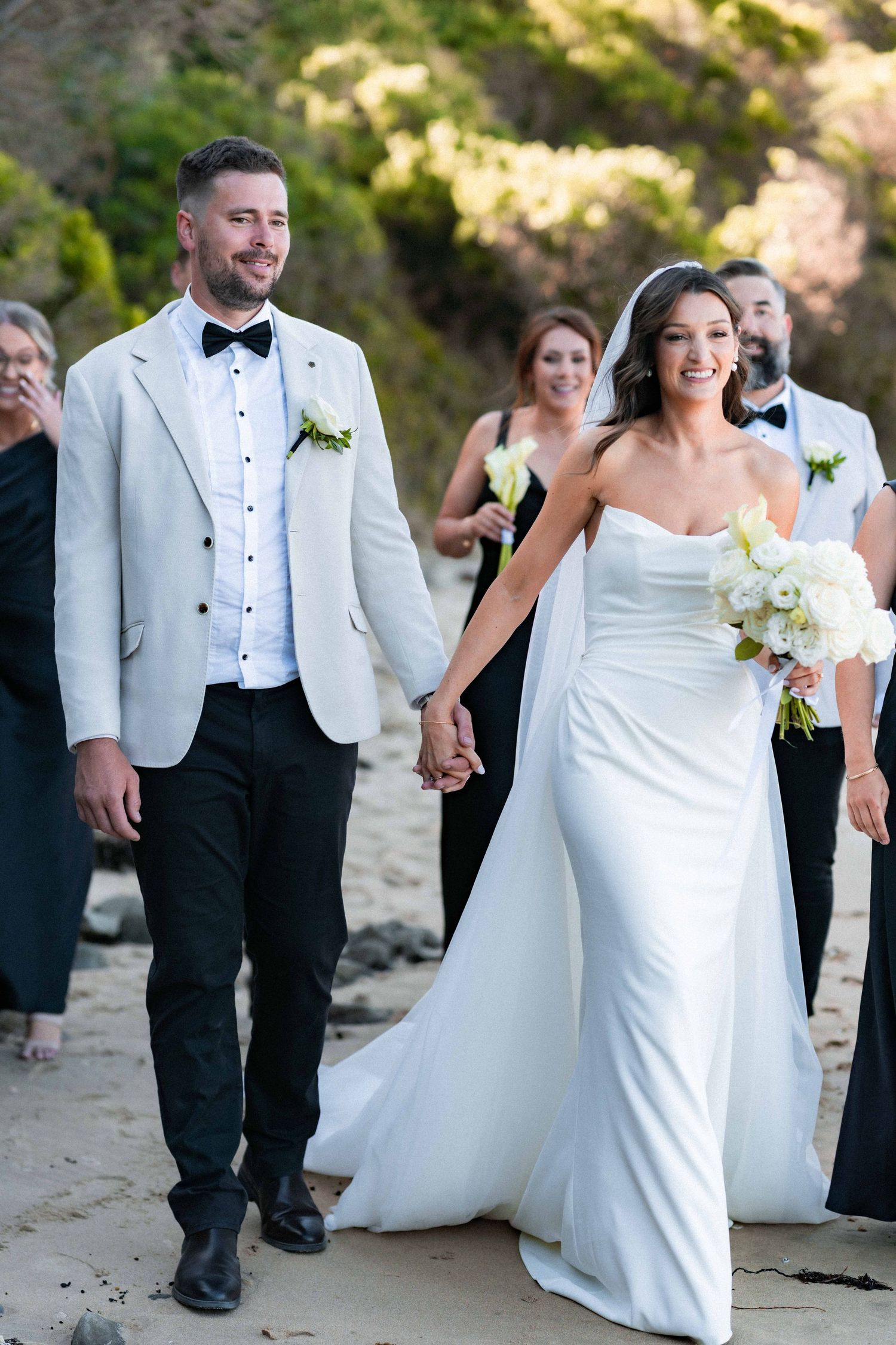A happy couple walks on the beach during their wedding ceremony, with guests following behind them.