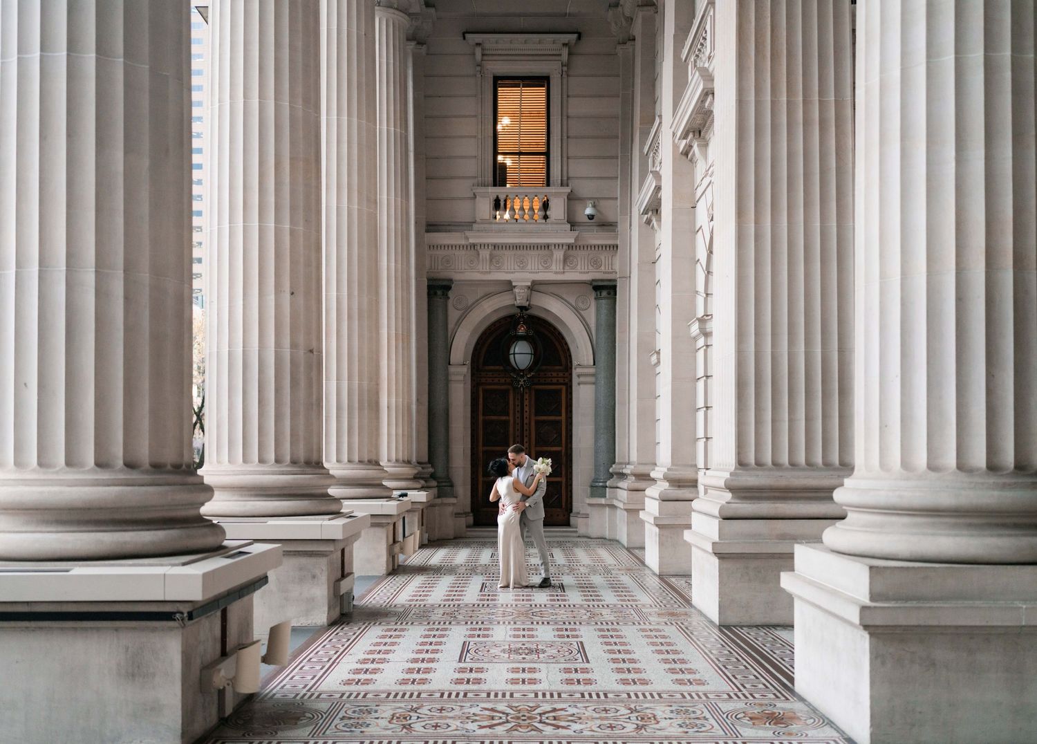 Couple embracing among grand white columns on ornate mosaic floor of a neoclassical building.