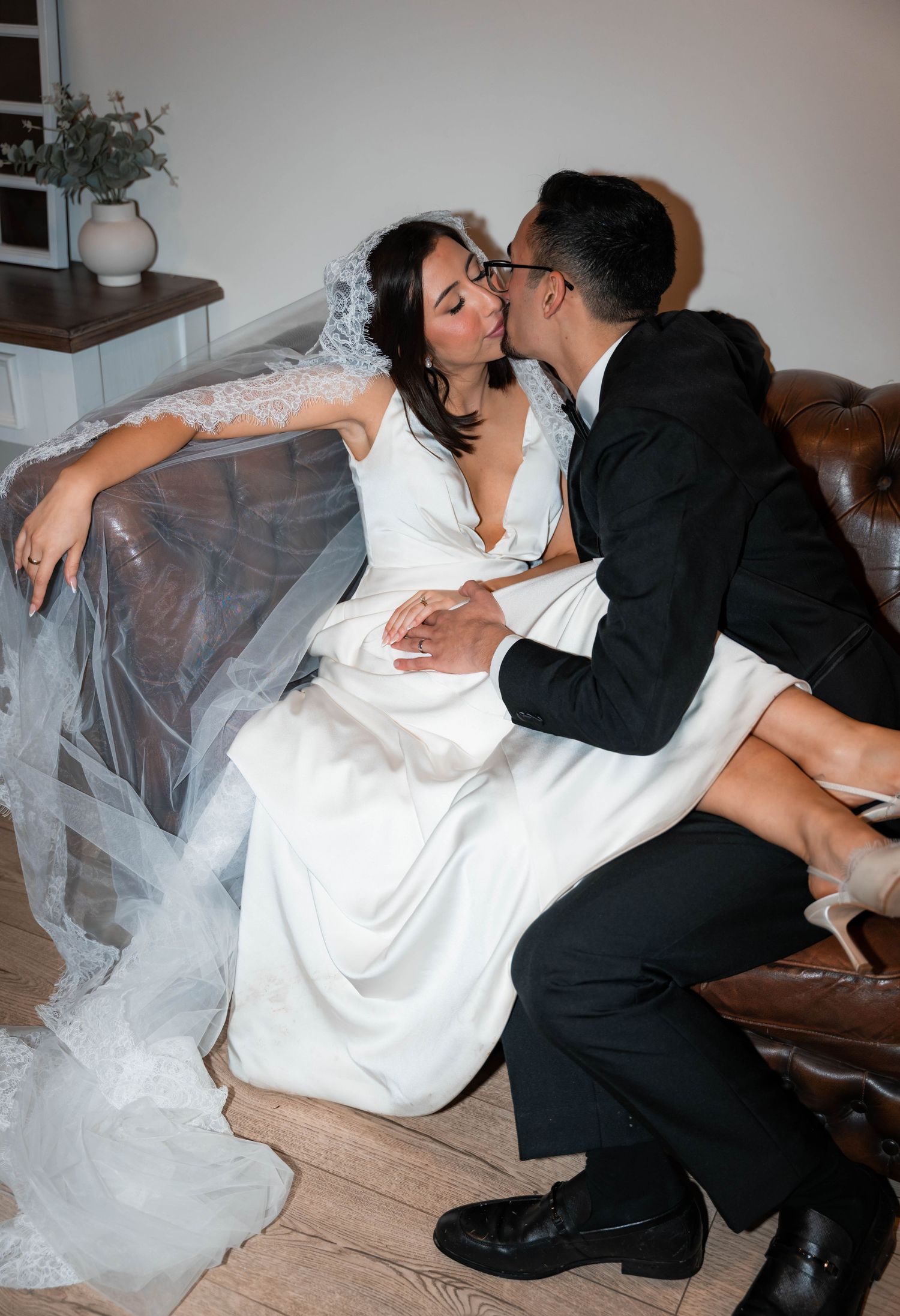 Romantic wedding couple sharing an intimate kiss while seated together in a beautifully decorated indoor setting.