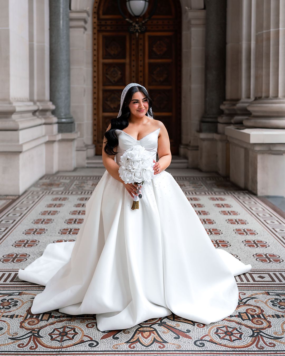 A bride in a flowing white wedding gown twirls on an ornate tiled floor in a grand historic building.