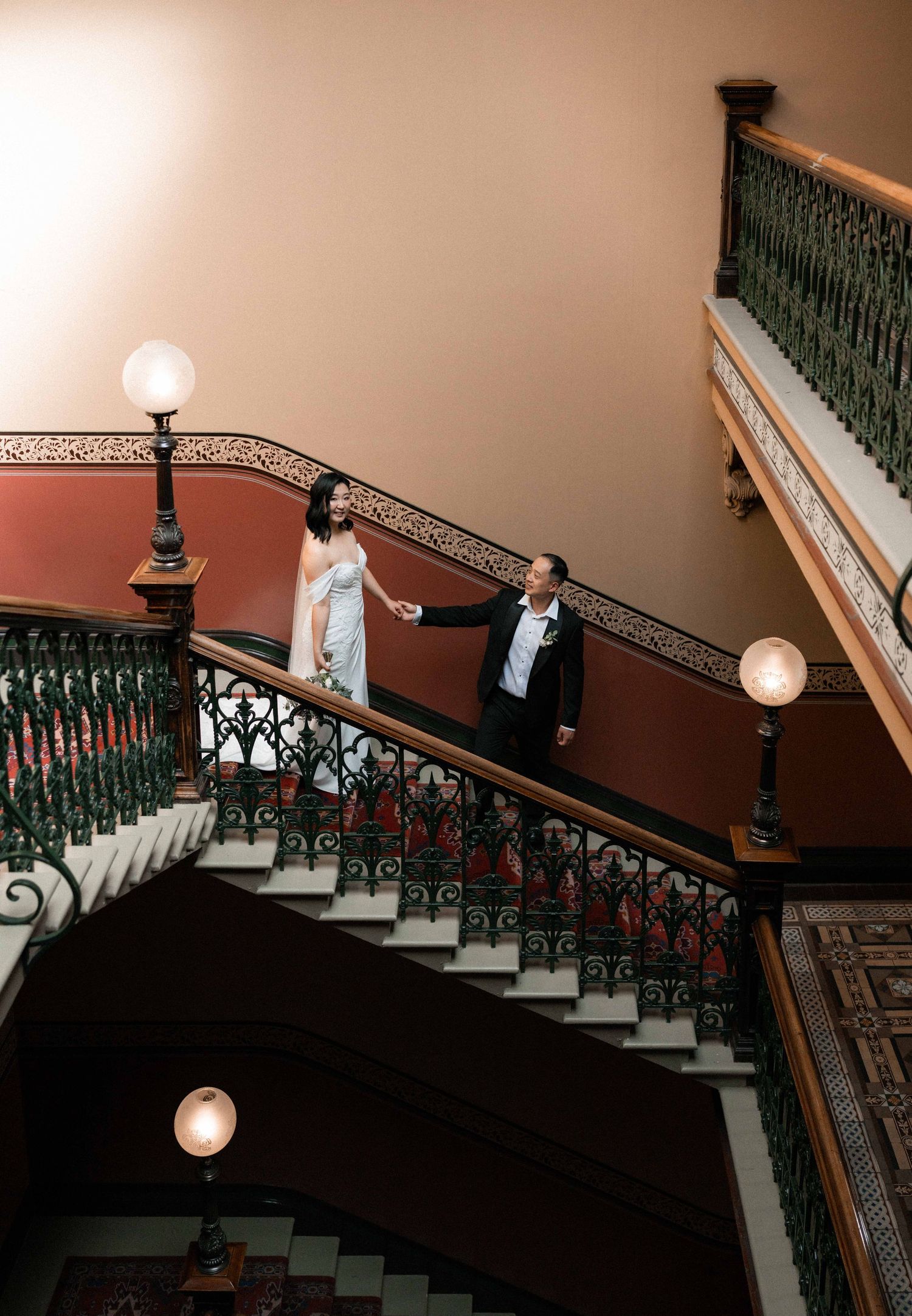 Couple in formal attire holding hands on ornate grand staircase with decorative iron railings and globe lights.