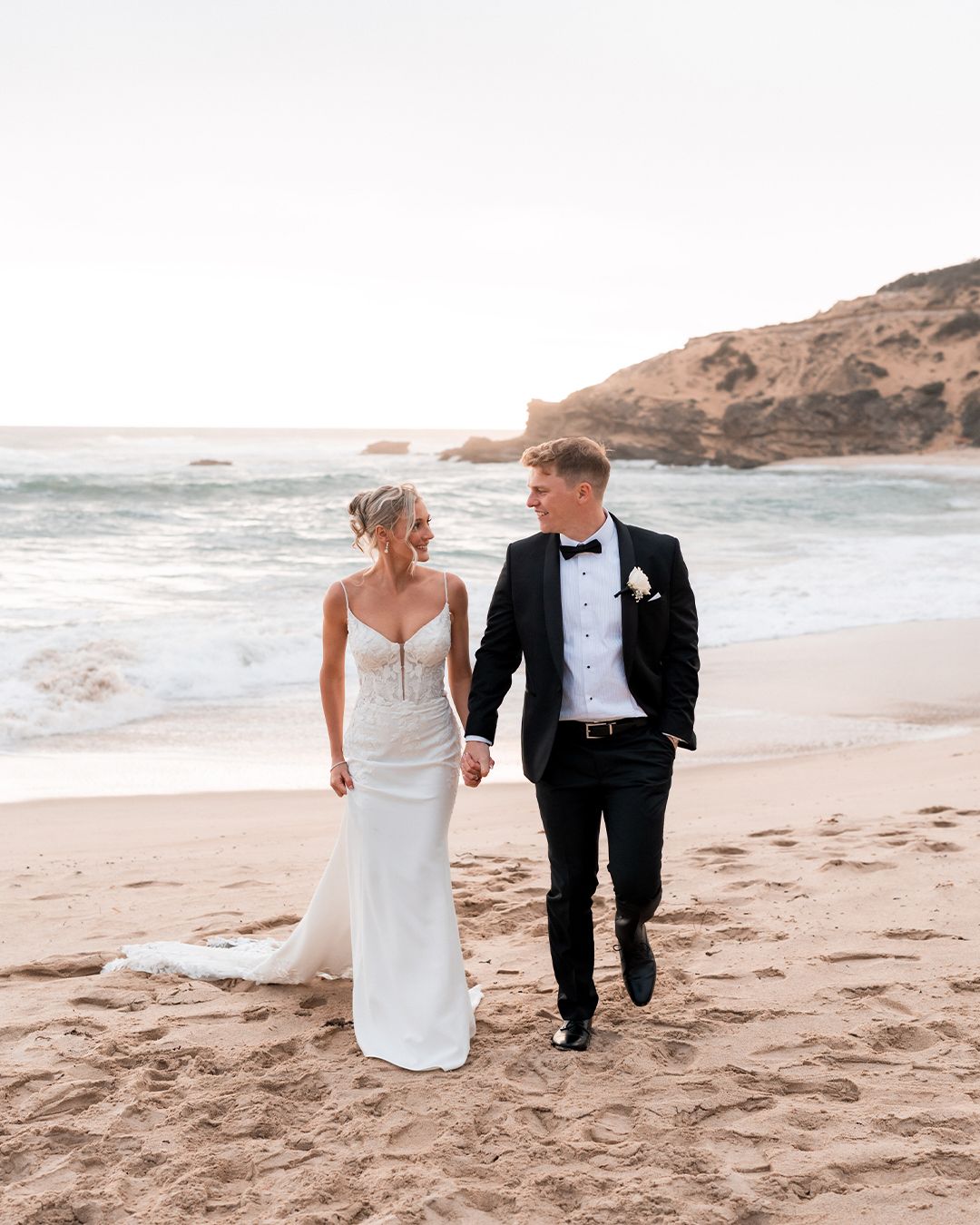Bride and groom walking hand in hand on a sandy beach at sunset, wearing wedding dress and black tuxedo.
