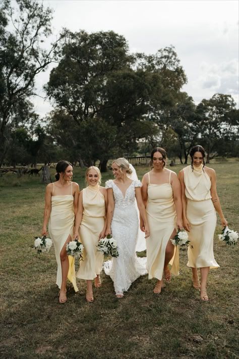 A bridal party walks together across a grassy field wearing cream-colored dresses and holding white floral bouquets.