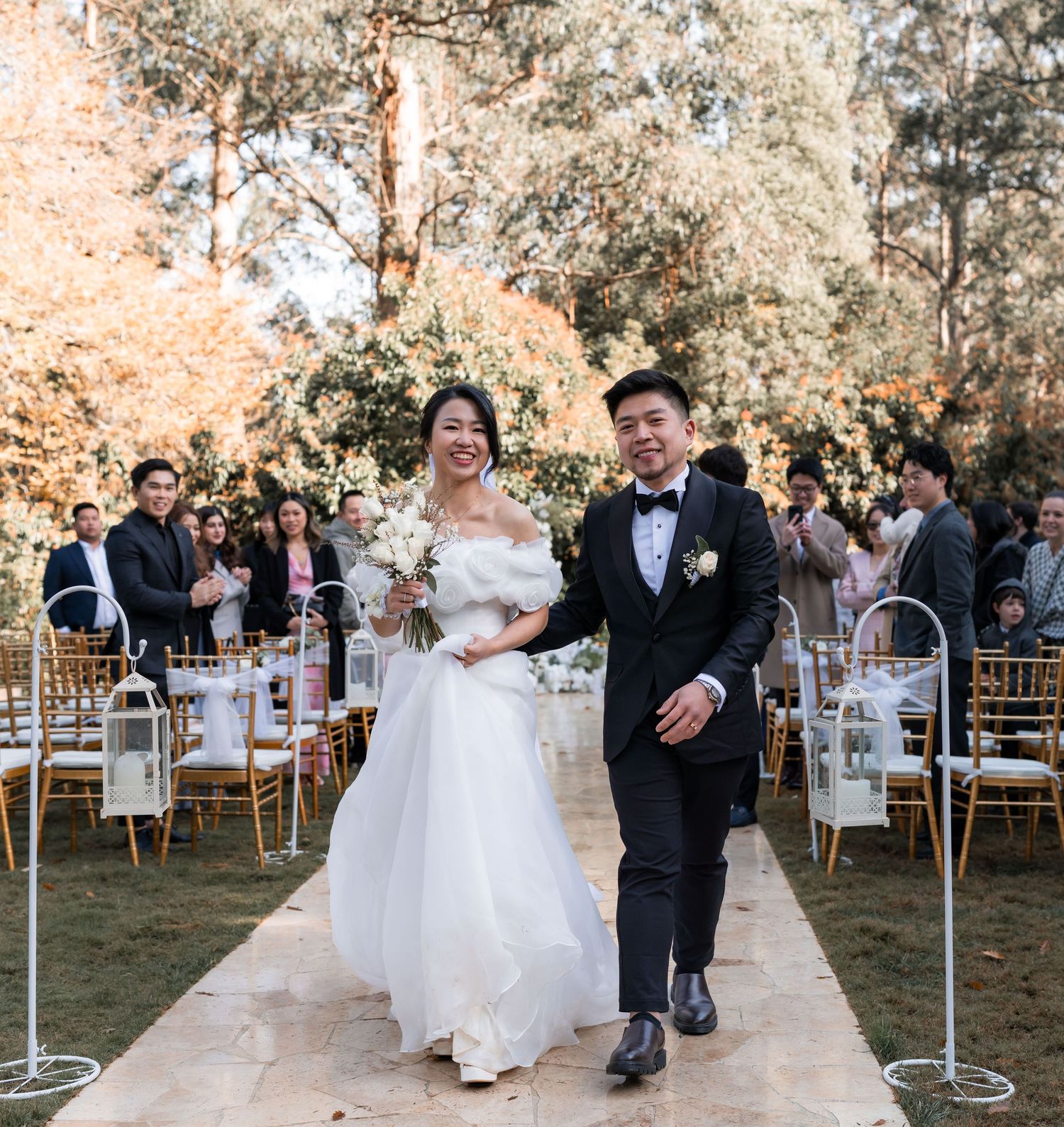 A couple walks down an outdoor aisle lined with white chairs and floral arrangements at a garden wedding ceremony.