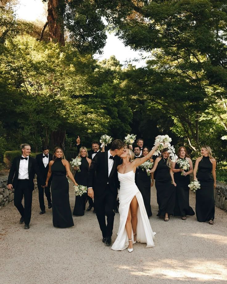 A wedding party in formal black attire and white dress walk together on a path surrounded by lush green trees.