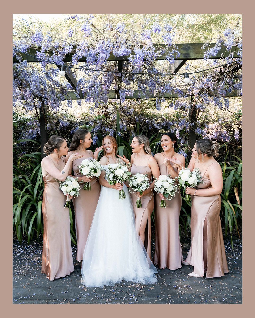 A bridal party in champagne-colored dresses poses together under blooming cherry blossom trees in spring.