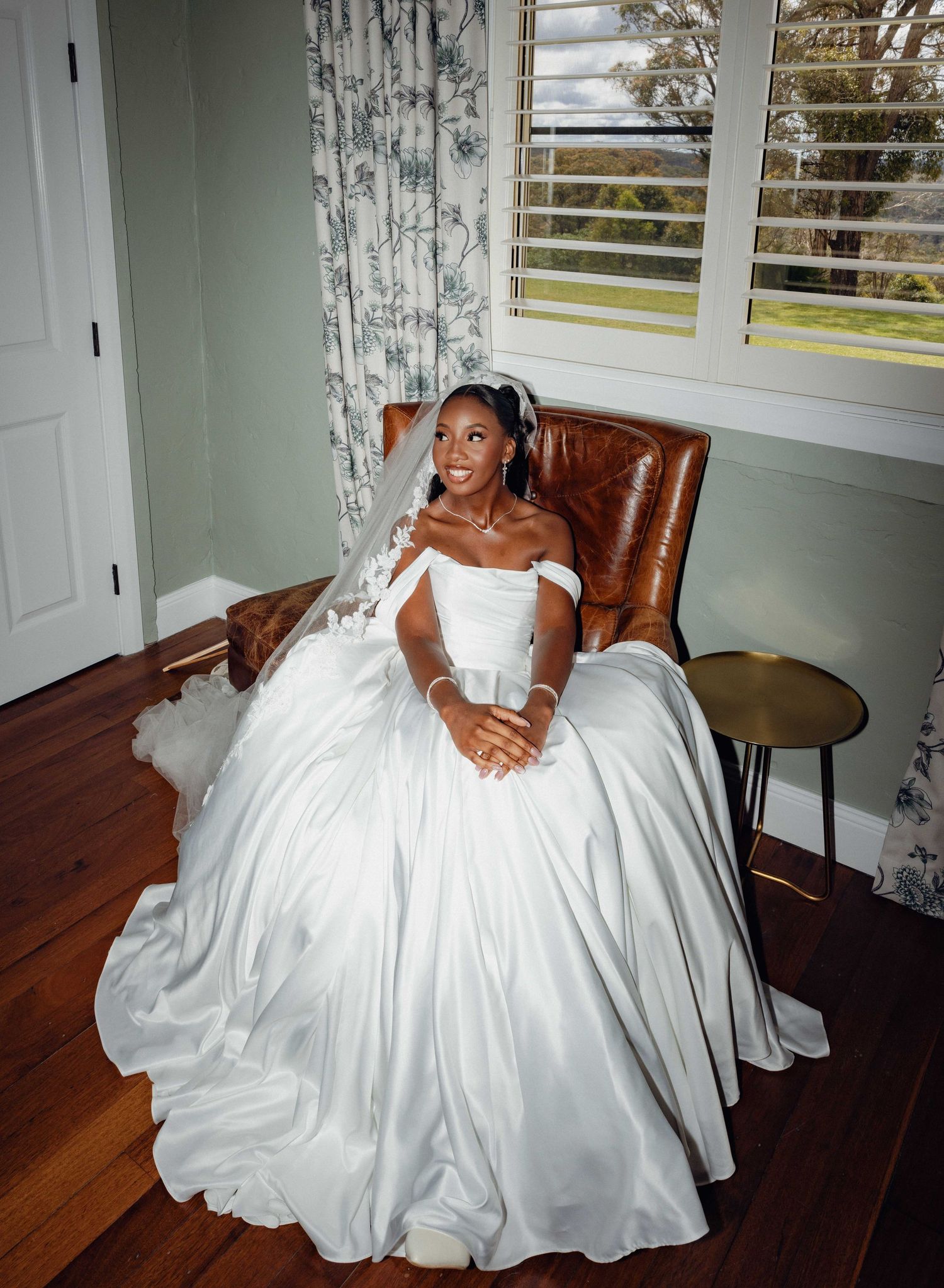 A person in an elegant white wedding gown poses gracefully in a brown leather armchair by a window.