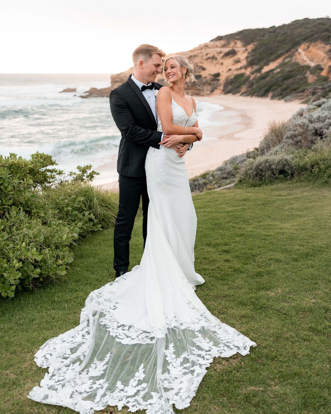 Bride in lace-train gown and groom in tuxedo embrace on coastal cliff with sandy beach backdrop.