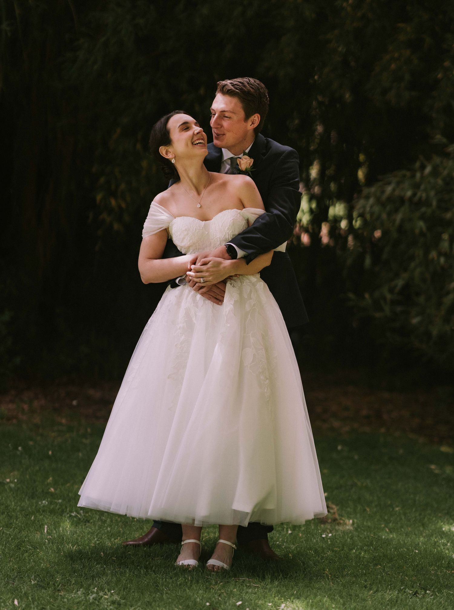 A romantic wedding moment captured outdoors with a bride in a tea-length tulle dress embracing with her partner.