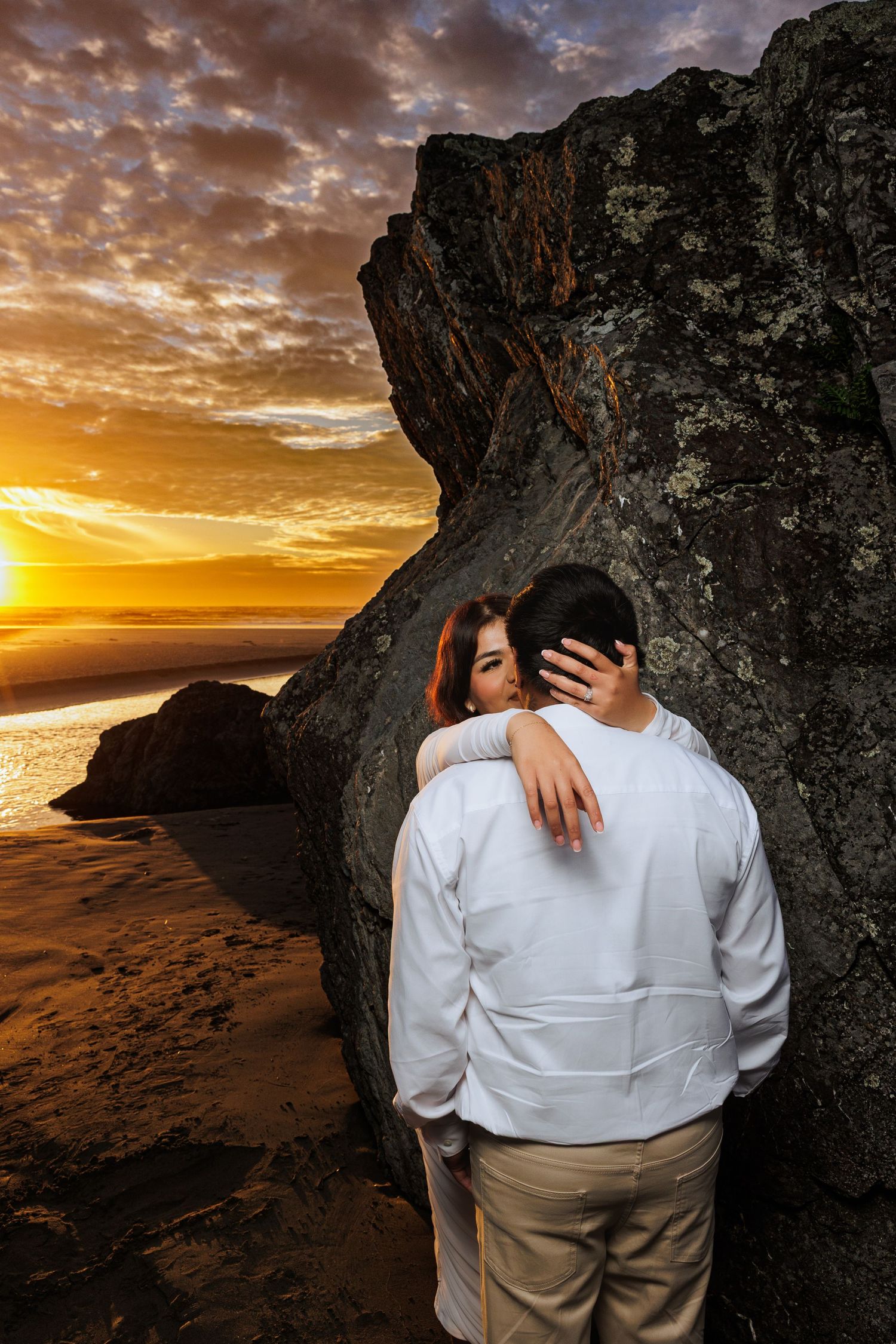 Couple embraces beside a coastal rock formation during a dramatic sunset portrait.