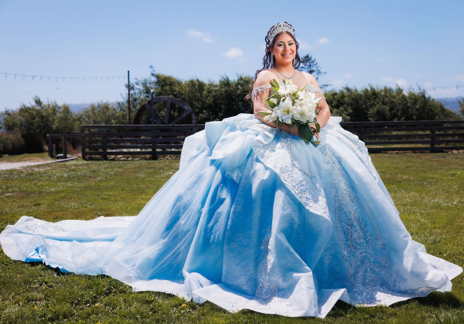 Quinceañera stands outdoors in a blue ball gown while holding a bouquet of white flowers.