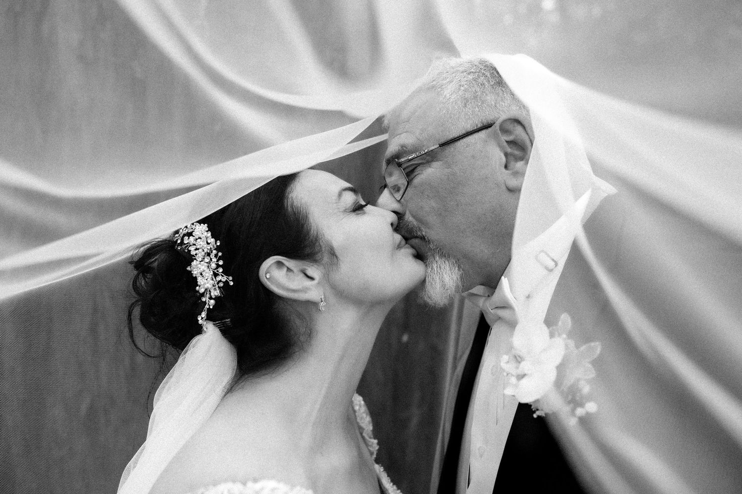 Bride and groom kiss beneath a flowing wedding veil in a black-and-white portrait.