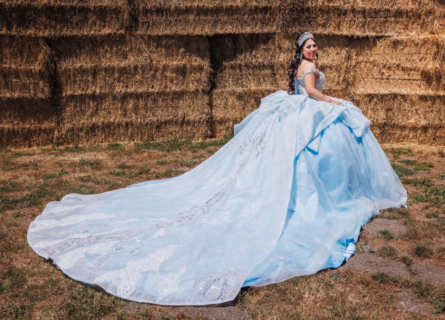 Quinceañera poses beside stacked hay bales with the long train of her blue gown spread across the ground.