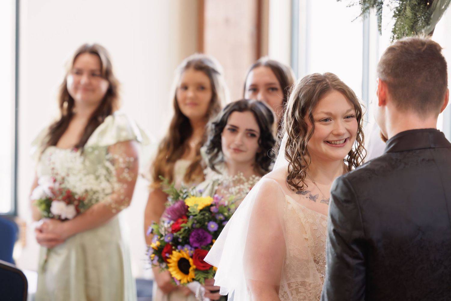 Smiling bride approaches the groom during an indoor wedding ceremony with her bridesmaids behind her.