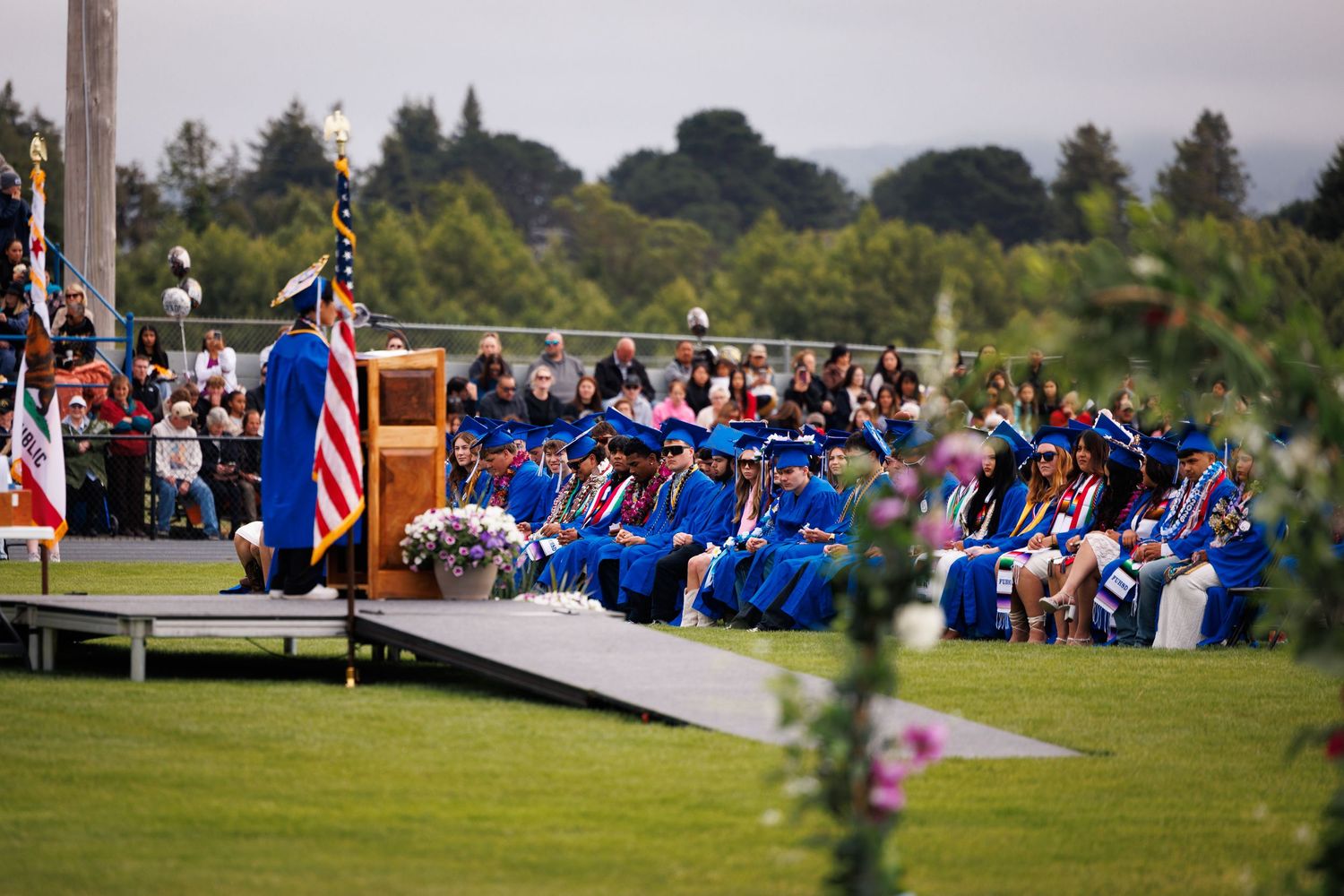 Graduating students in blue caps and gowns sit together during an outdoor graduation ceremony.