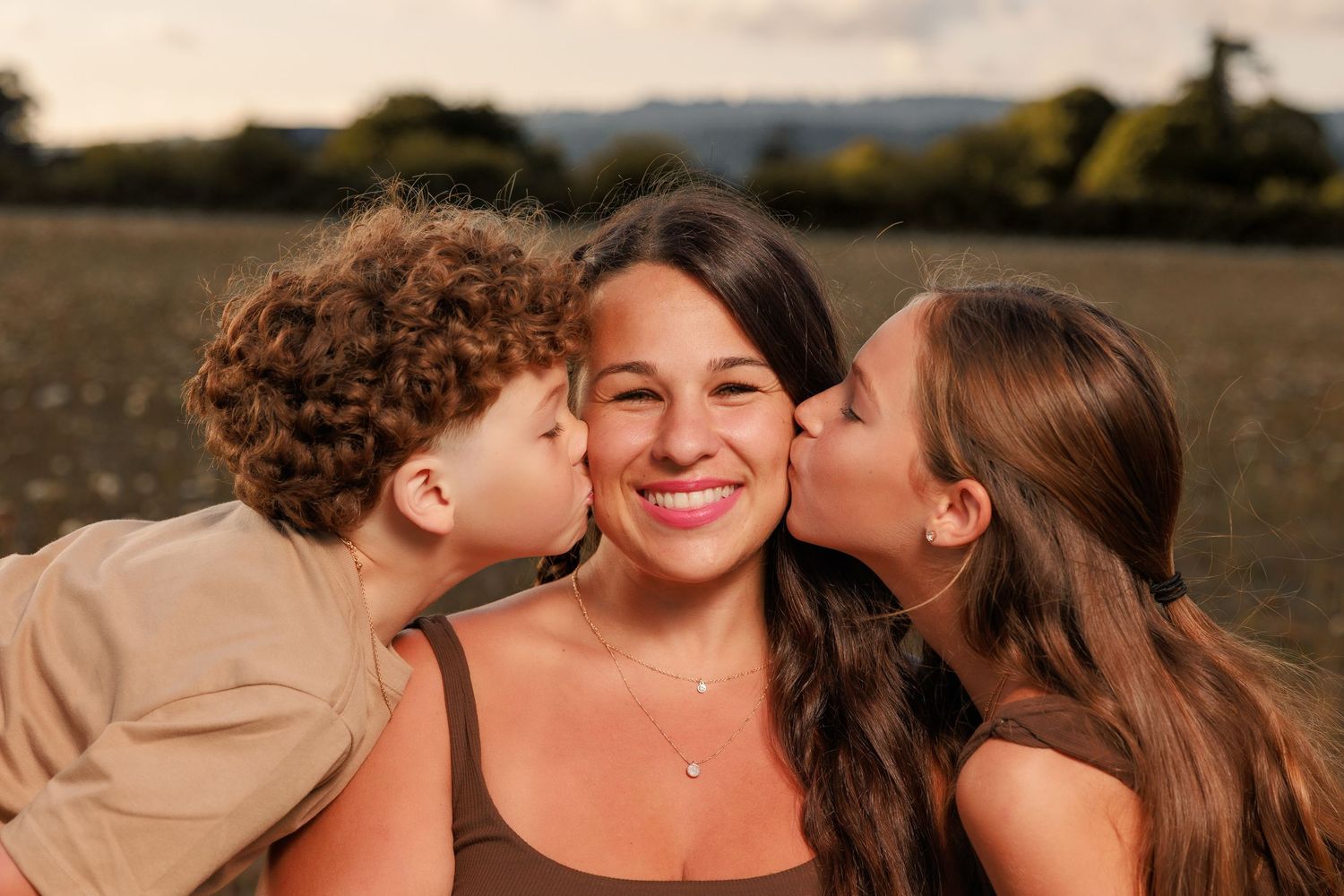 Mother smiling as her two children kiss her cheeks during a warm outdoor family portrait session.