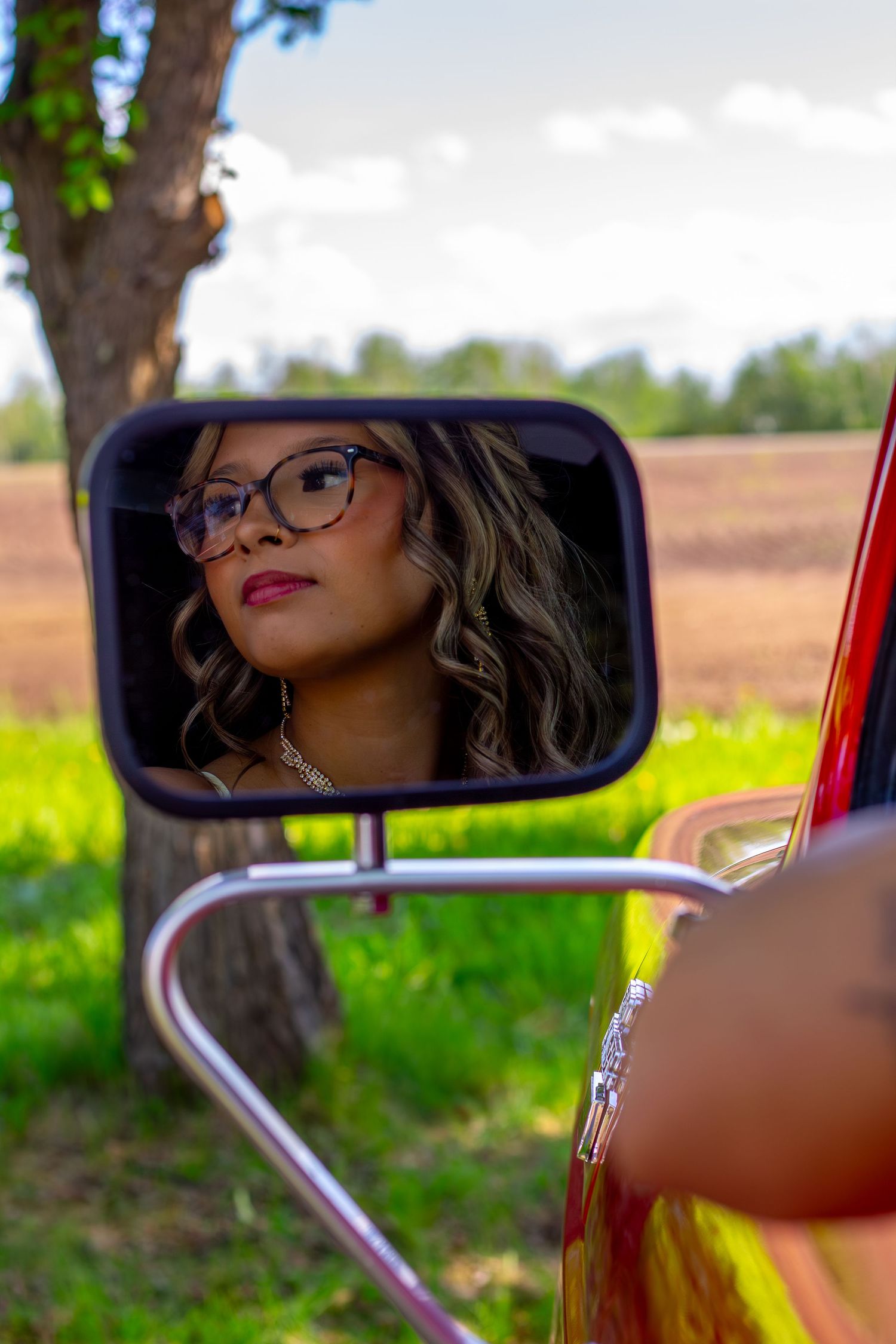 Young woman with glasses and curly hair reflected in a vintage red truck side mirror outdoors.