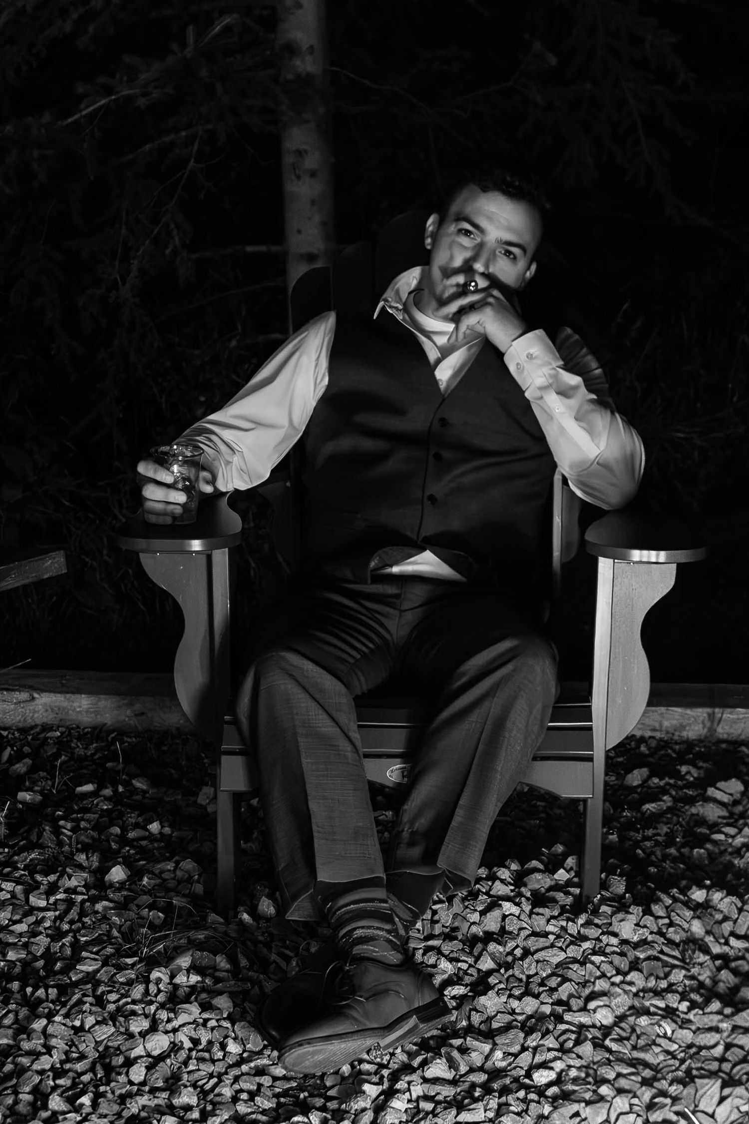 Black and white portrait of a man in a vest sitting in a wooden chair, smoking a cigar and holding a glass.