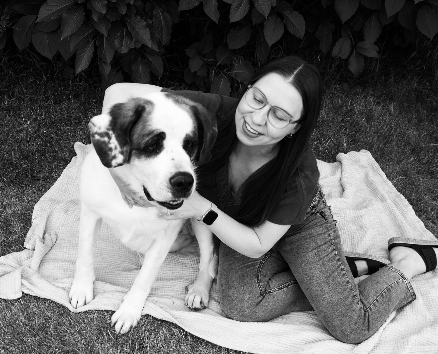 Black and white photo of smiling woman with glasses hugging large dog on blanket.