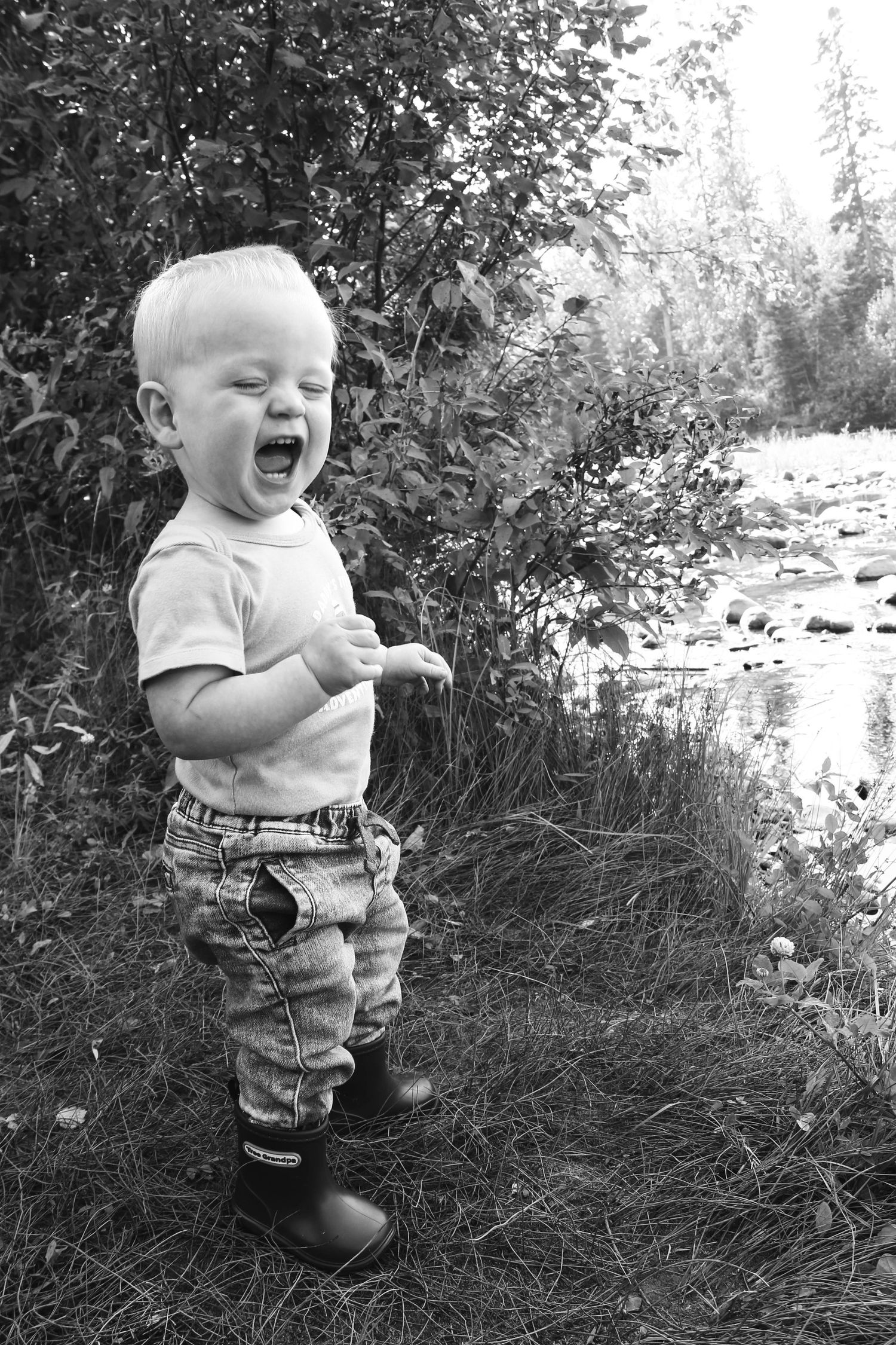 Laughing toddler boy standing near creek in black and white outdoor portrait photo.