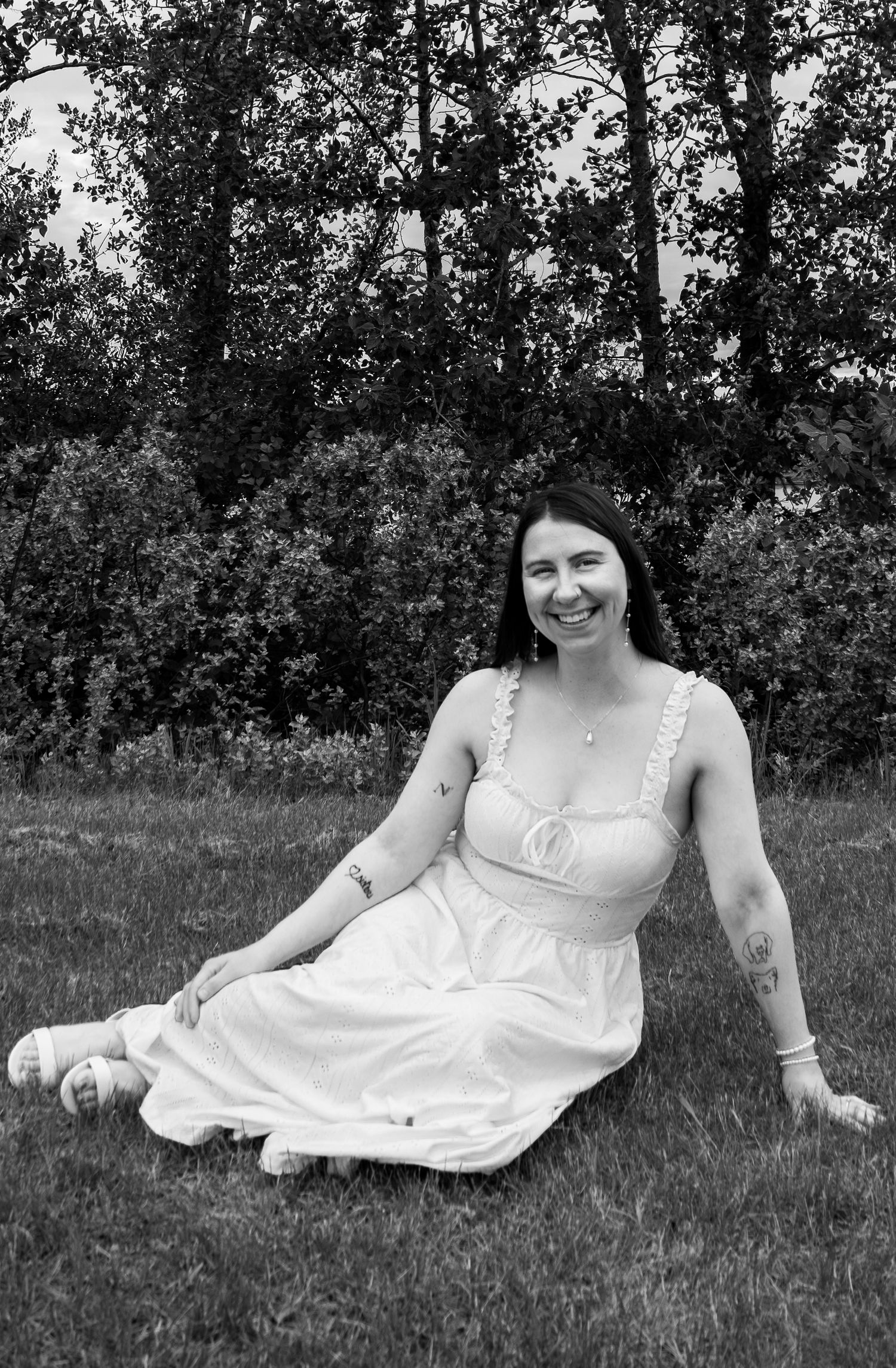 Woman in white sundress sitting on grass smiling in a black and white outdoor portrait photo.