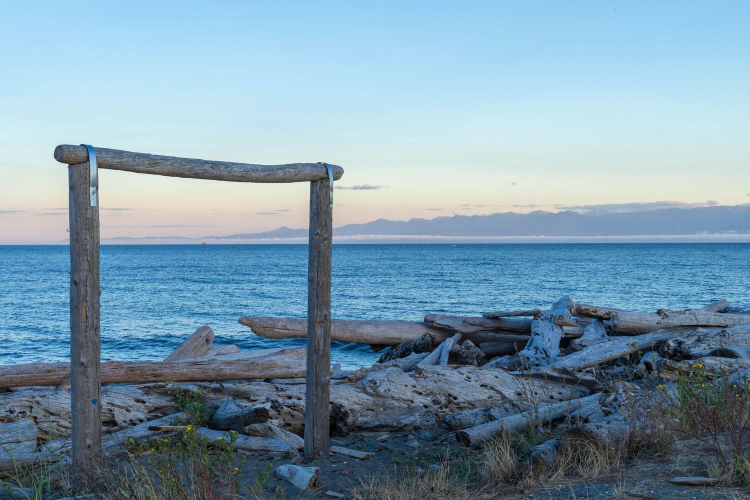 Rustic driftwood frame structure on rocky beach at sunset with calm ocean and mountains.