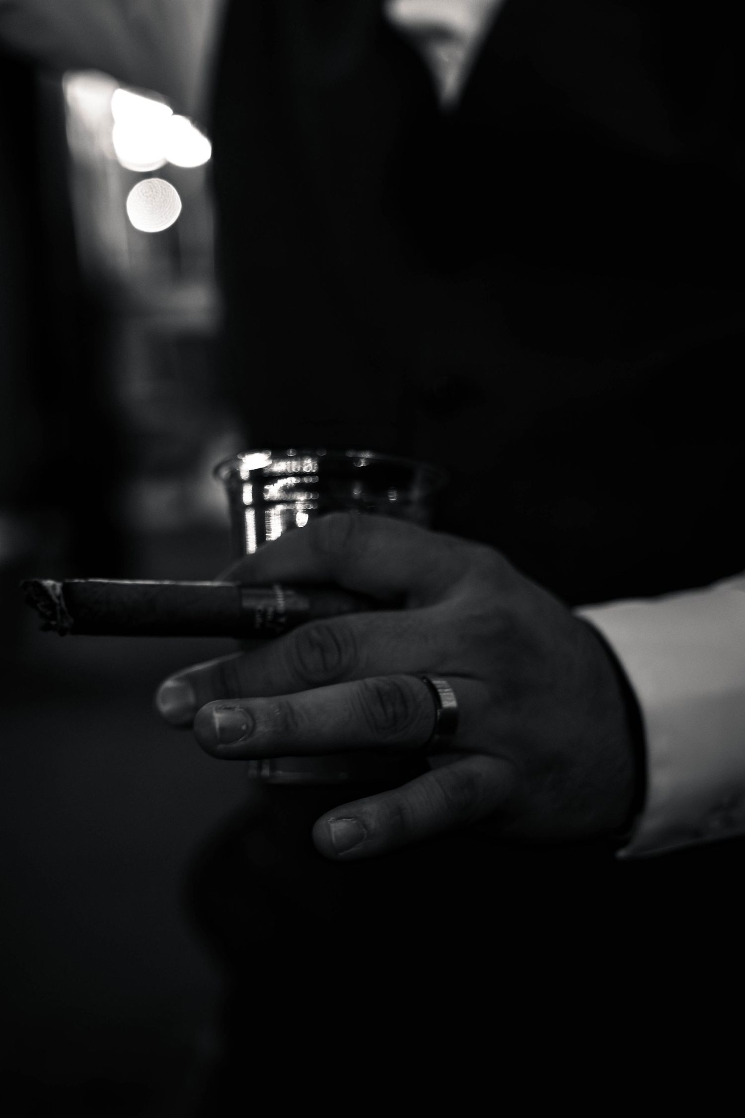 Black and white photo of a mans hand holding a cigar and glass, wearing a ring, in a dimly lit setting.