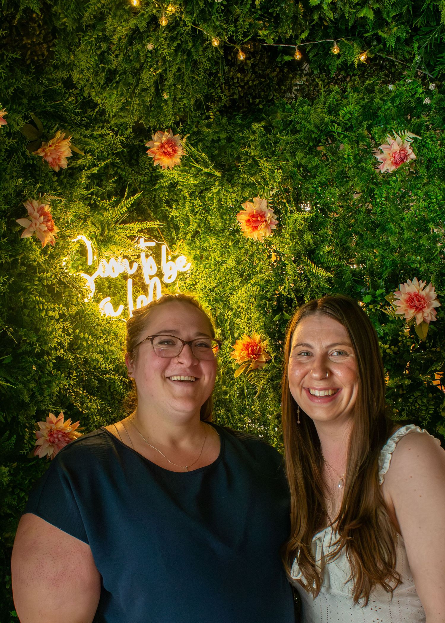 Two smiling women posing in front of green floral wall with glowing neon sign decor.