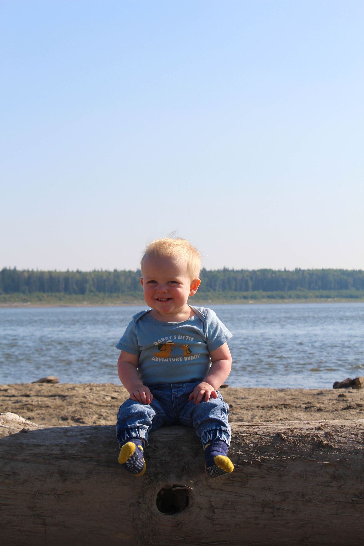 Happy smiling baby boy sitting on log at lakeside on sunny summer day.