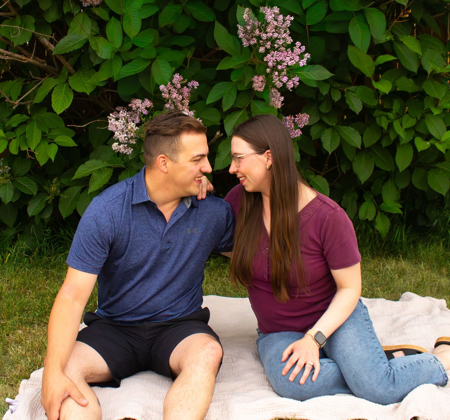 Happy couple sitting on blanket laughing together in front of blooming lilac bushes.