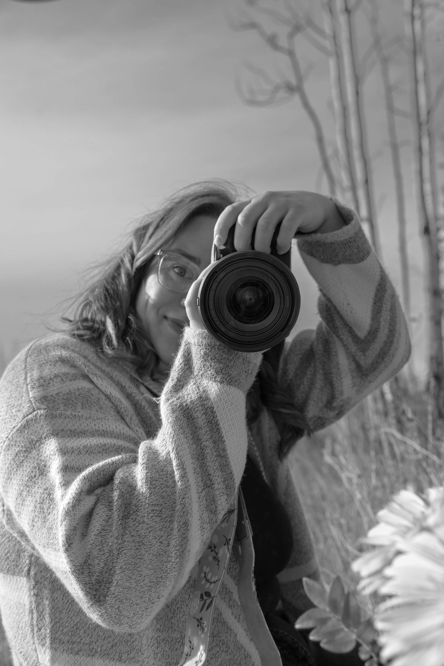 Black and white photo of a young woman holding a DSLR camera up to her face outdoors near bare trees.