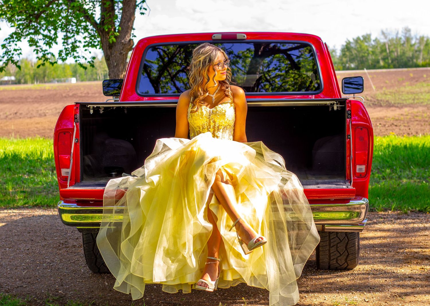 Young woman in a flowing yellow prom dress sitting on the tailgate of a red pickup truck outdoors.